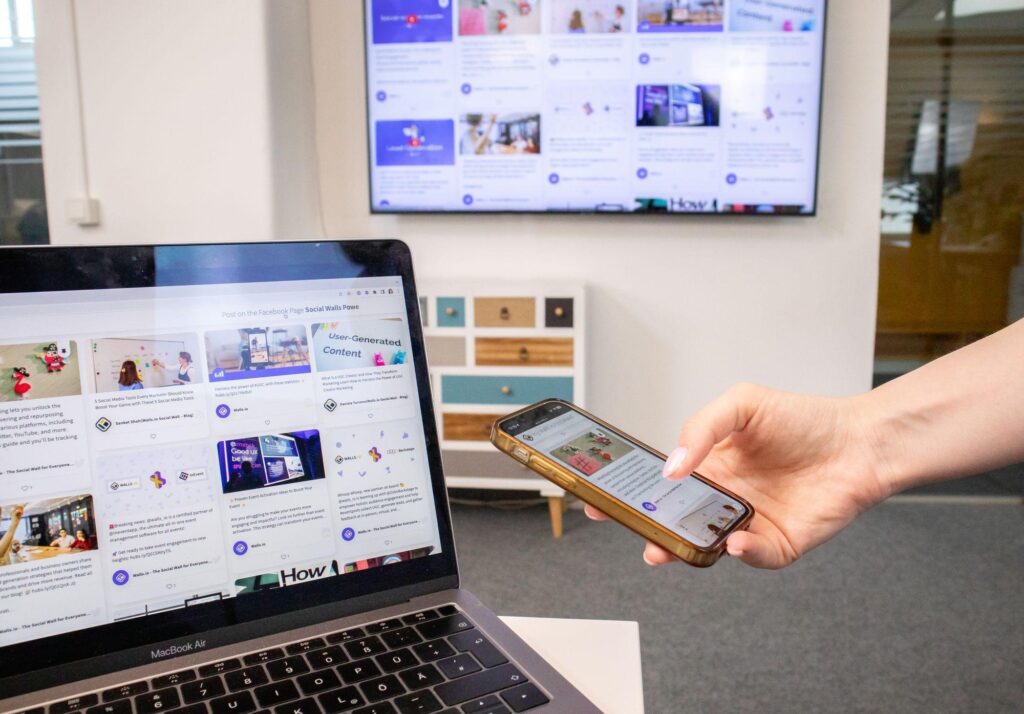 Hand holding a phone showing a social media dashboard, with a laptop and screen in the background